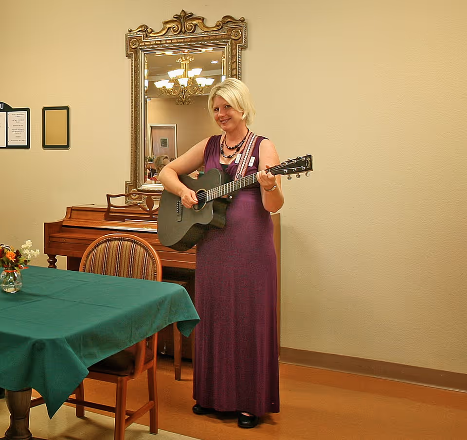A woman in a long purple dress playing a black acoustic guitar in a room with a green tablecloth-covered table, a wooden chair, a piano, and a large ornate mirror on the wall.
