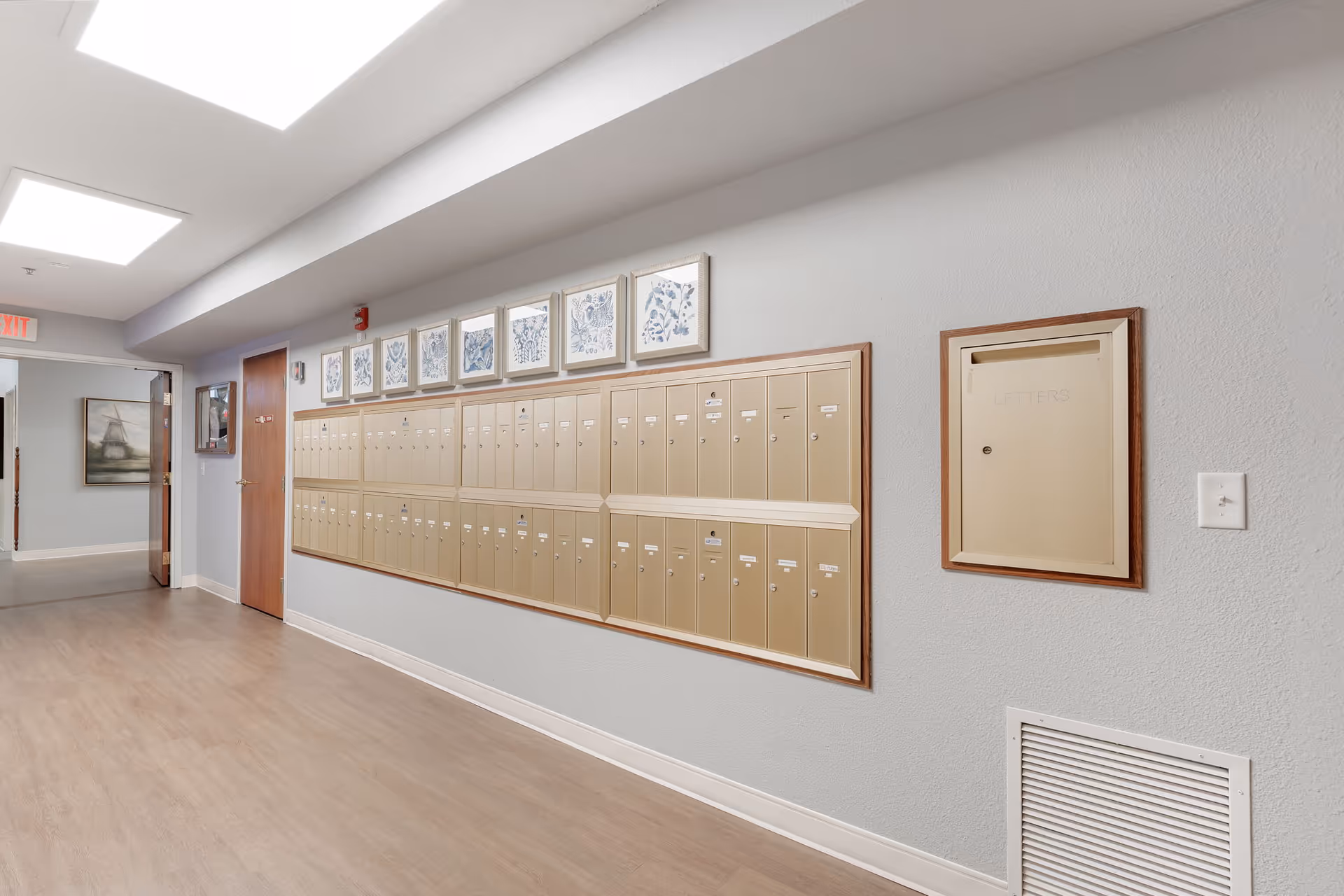 Interior hallway with a row of beige mailboxes mounted on a light gray wall. Above the mailboxes are framed blue and white artwork. The floor is wood-style laminate, and there is a door and an exit sign visible at the end of the hallway.