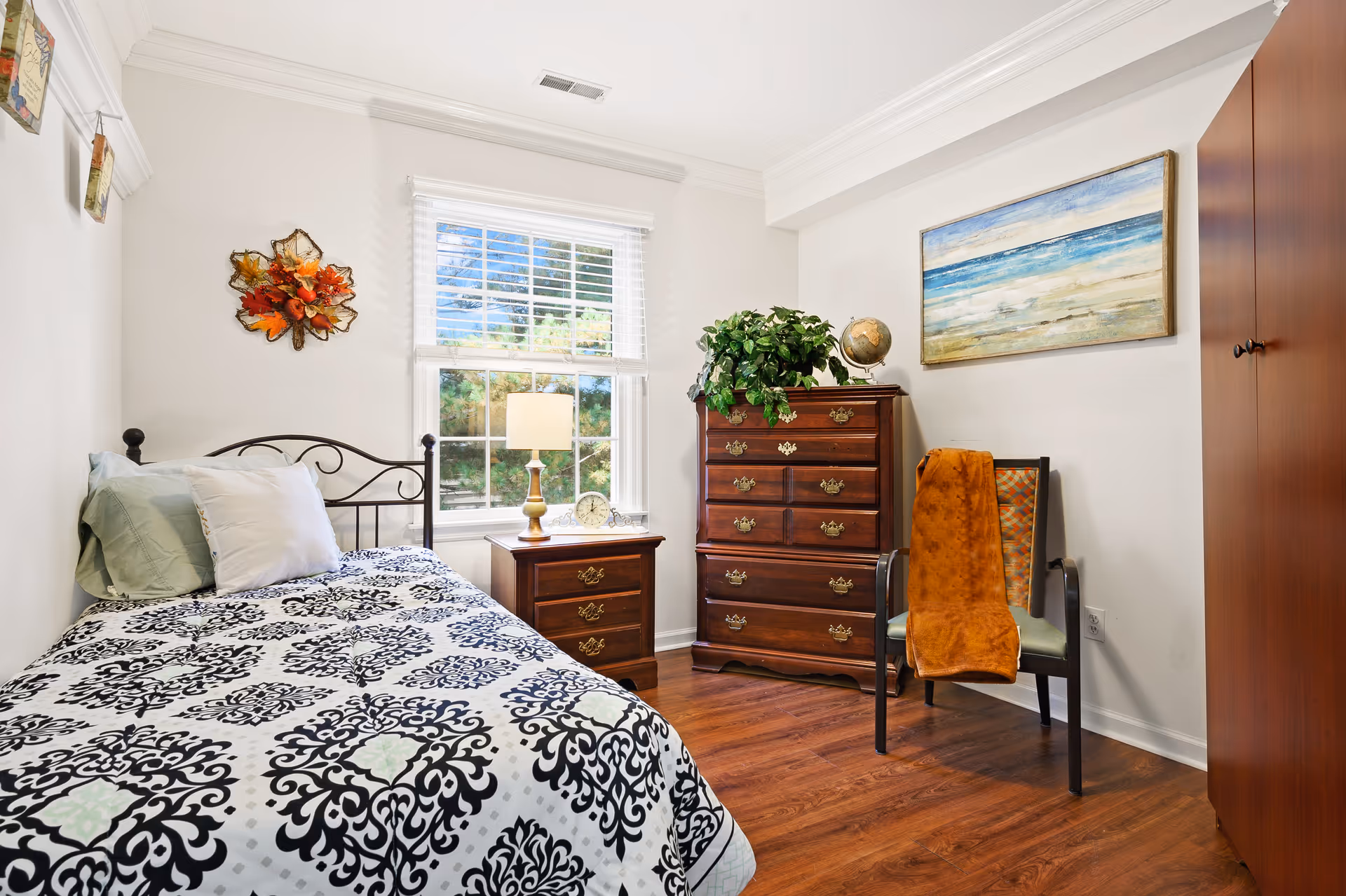 A cozy bedroom with a single bed covered in a black and white patterned bedspread, two pillows, and a metal headboard. Next to the bed is a wooden nightstand with a lamp and a clock. A window with white blinds lets in natural light. Across from the bed is a wooden dresser with brass handles, topped with a green plant and a globe. A chair with an orange blanket draped over it sits beside the dresser. A large painting of a beach scene hangs on the wall above the chair. The room has wooden flooring and white walls.