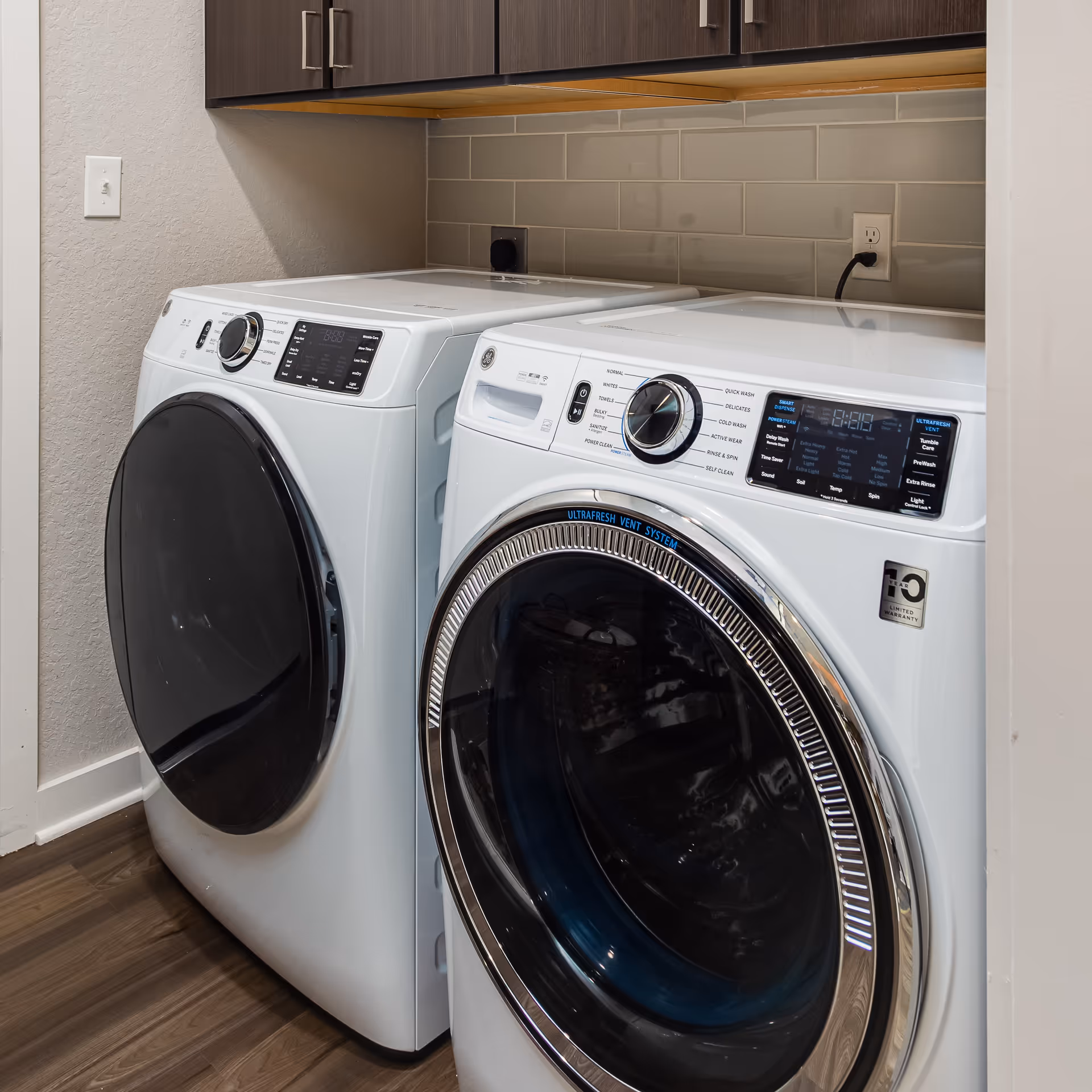 A modern laundry area with a white front-loading washing machine and dryer side by side, set against a wall with gray subway tile backsplash and dark wooden cabinets above.