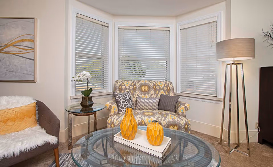 A cozy living room corner with a patterned armchair adorned with three decorative pillows, a glass coffee table with two yellow vases on a white tray, a side table with a white orchid plant, a modern floor lamp, and a framed abstract painting on the wall. The room has three windows with white blinds.