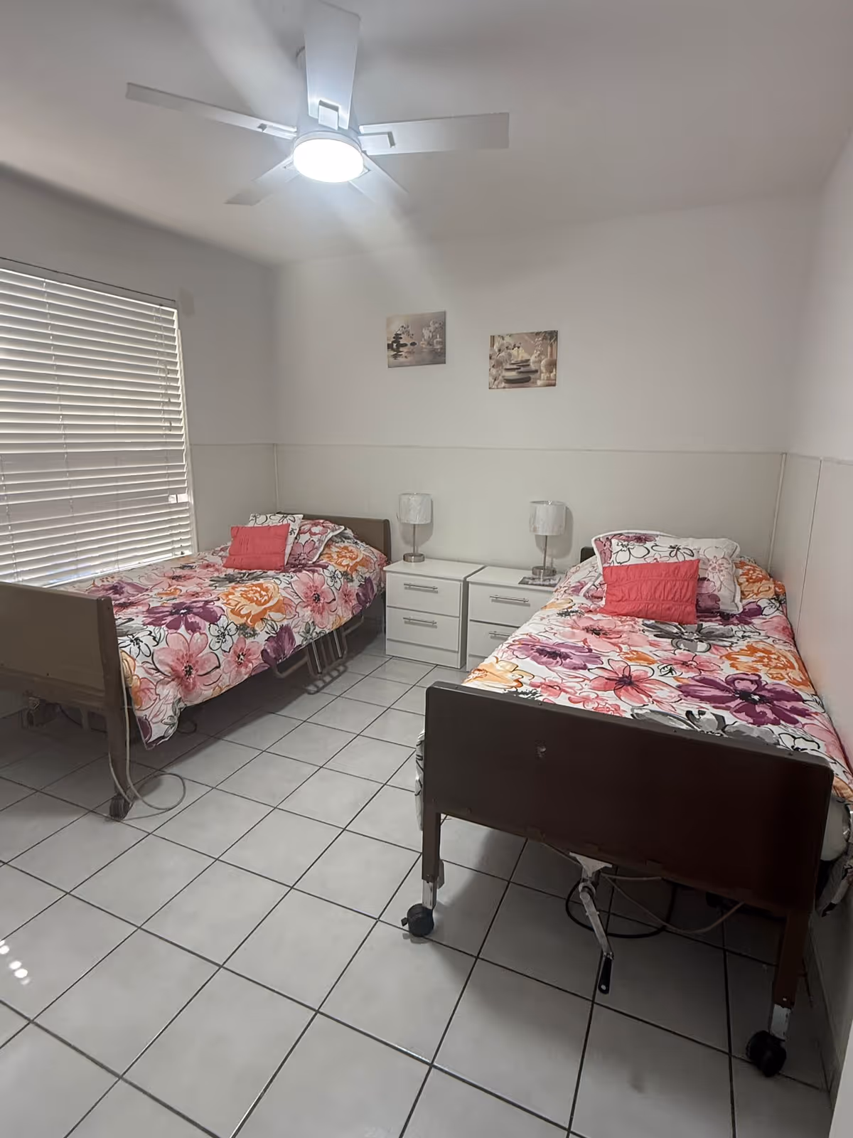 A small bedroom with two single hospital-style beds on wheels, each covered with floral bedspreads in shades of pink, purple, and orange. Between the beds are two white nightstands with two matching lamps. The room has white tiled floors, white walls, a window with closed blinds, and a ceiling fan with a light.
