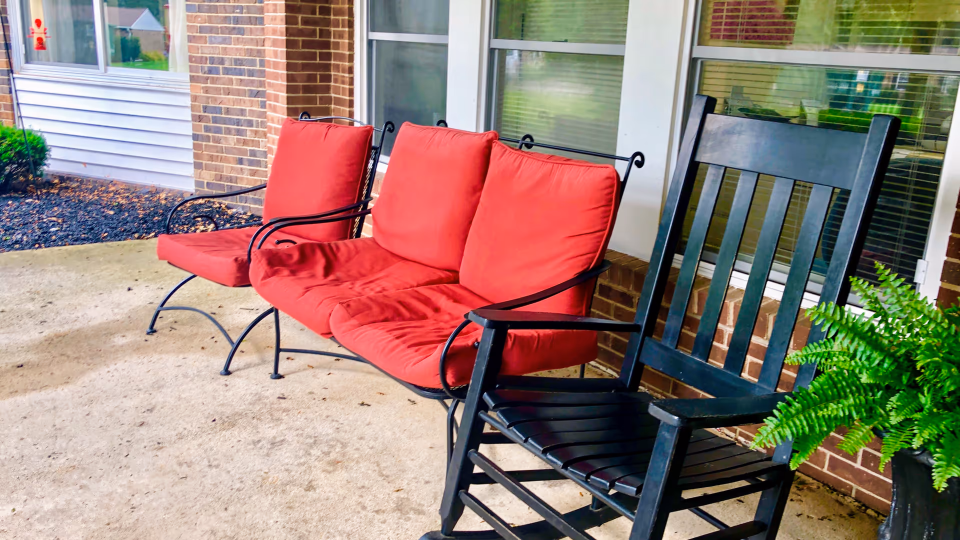 Red-cushioned metal loveseat and black wooden rocking chair on a covered brick porch with windows and a potted fern.