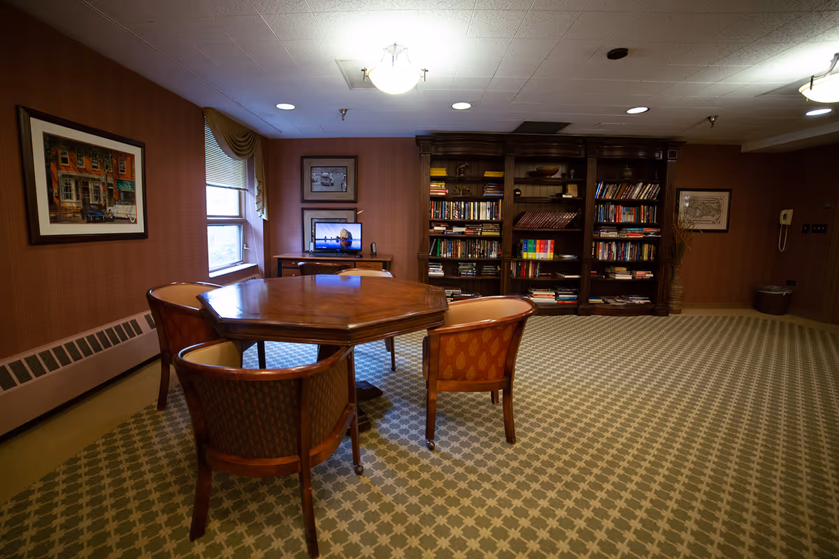 A cozy interior room with a wooden octagonal table surrounded by four upholstered chairs on a patterned carpet. Against the back wall is a large wooden bookshelf filled with books and decorative items. A small desk with a computer monitor is positioned near a window with draped curtains. The walls are adorned with framed pictures, and the ceiling has recessed lighting and a central light fixture.