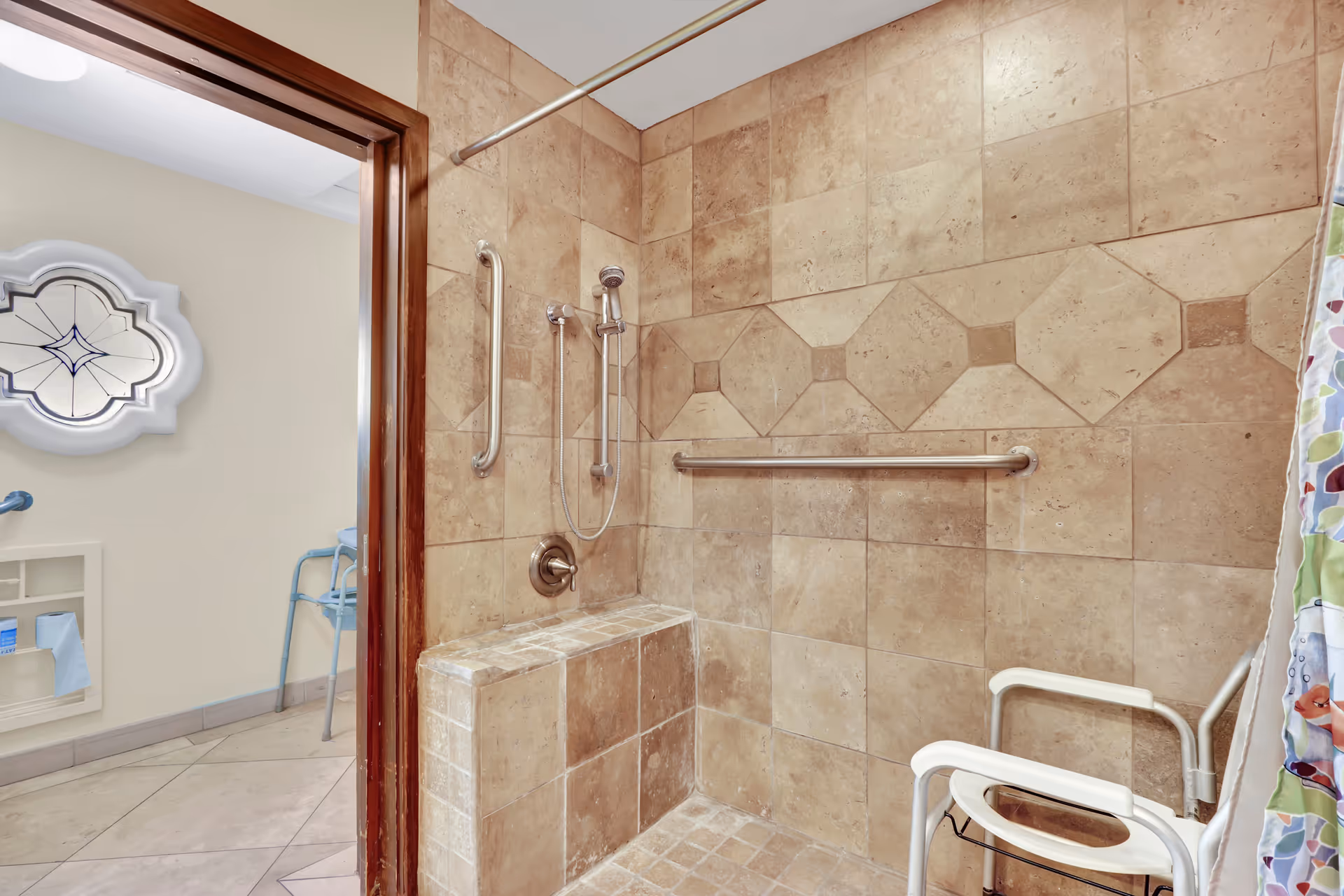 A tiled shower area with beige stone tiles, a built-in bench, a handheld showerhead, and two metal grab bars. A white shower chair is positioned inside the shower. The shower curtain has a colorful pattern. The bathroom door is open, showing a beige wall with a decorative window and a blue walker in the adjacent room.