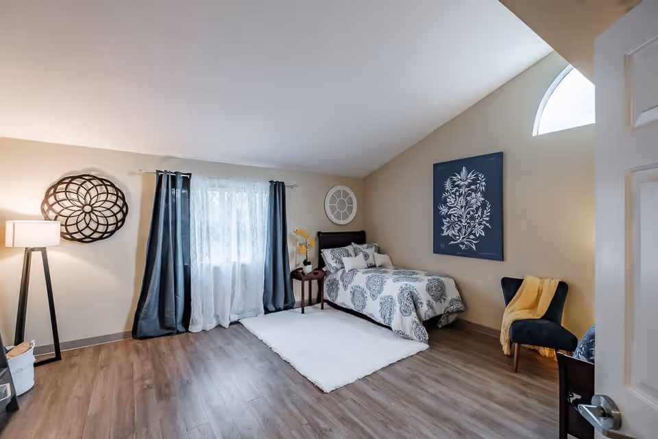 A cozy bedroom with a single bed covered in a white and gray patterned comforter. The room features a white rug on a wooden floor, a small side table with a vase of yellow flowers, a dark upholstered chair with a yellow throw blanket, and decorative wall art. A window with sheer white curtains and dark drapes lets in natural light, and a floor lamp stands in the corner.