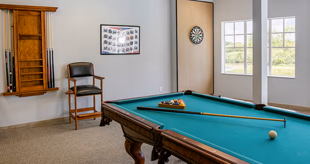 A recreational room with a pool table set up for a game, a cue stick resting on the table, a dartboard mounted on the wall, a chair, and a wooden rack holding pool cues. There are two large windows letting in natural light and a framed picture on the wall.