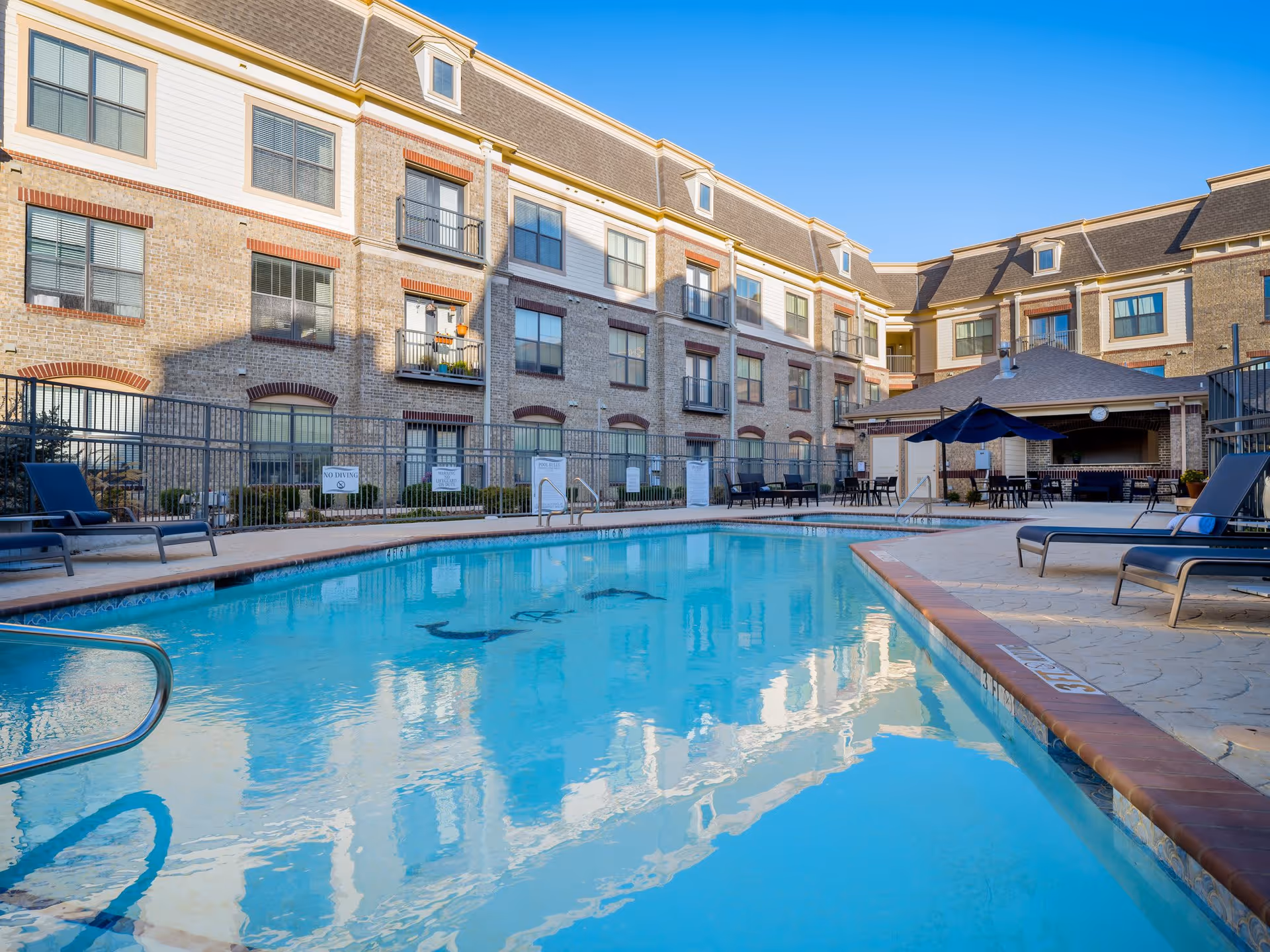 Outdoor swimming pool area at Discovery Village At Castle Hills with lounge chairs, tables with umbrellas, and a multi-story brick building in the background under a clear blue sky.