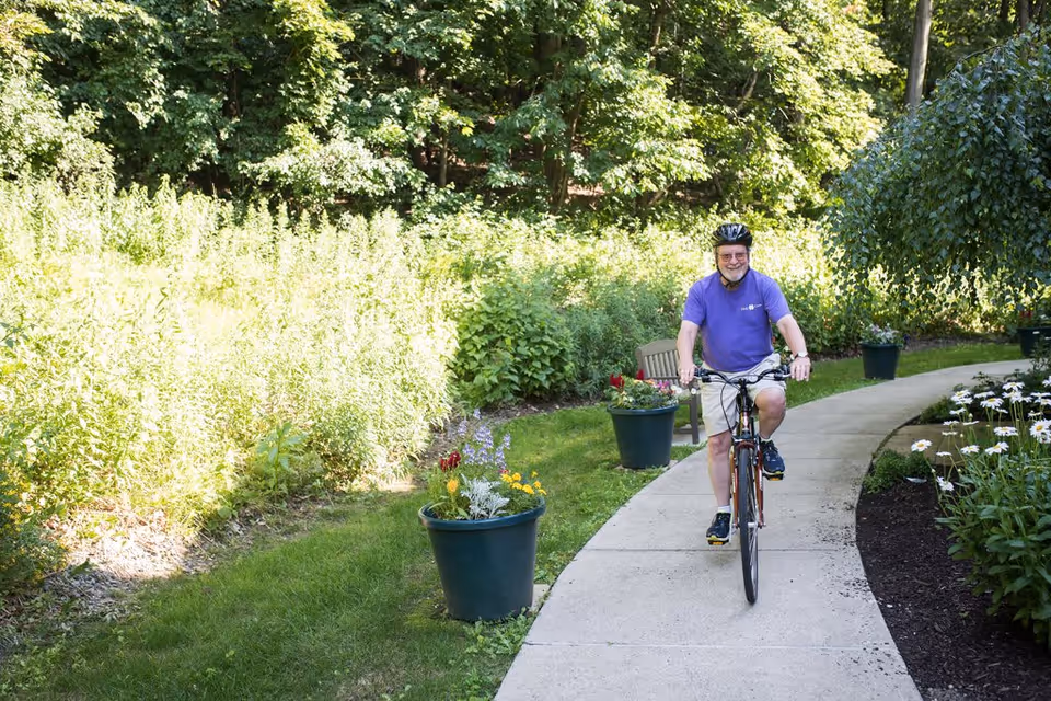 An elderly man wearing a purple shirt, beige shorts, and a black helmet is riding a bicycle on a curved concrete path surrounded by green plants, flowers, and trees in a garden setting.