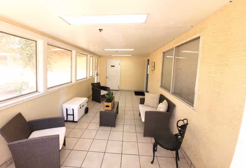 Narrow enclosed seating area with tiled floor, wicker chairs, small tables and windows along one side.