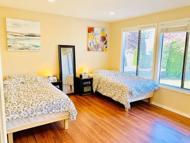 Bright bedroom with two twin beds, patterned bedding, bedside tables with lamps, a standing mirror and large window overlooking greenery.