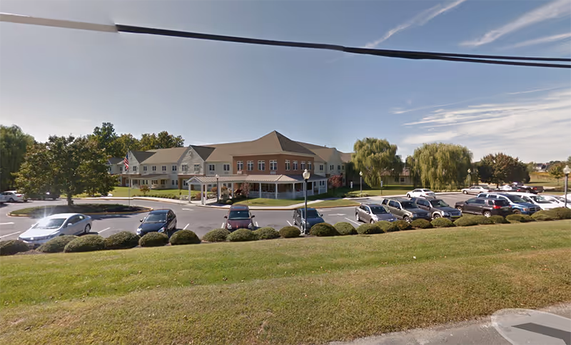 Exterior view of a senior living facility named Terraces At Parke Place with a large parking lot in front, surrounded by green grass and trees under a clear blue sky.