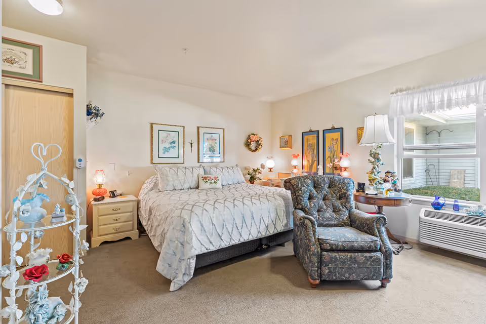 A cozy bedroom in an assisted living facility featuring a large bed with a patterned bedspread and multiple pillows. There is a floral upholstered armchair in front of the bed, a wooden nightstand with a lamp, and decorative framed artwork on the walls. A window with sheer curtains lets in natural light, and a small table with a lamp and decorative items is placed near the window. The room has carpeted flooring and a light, neutral color scheme.