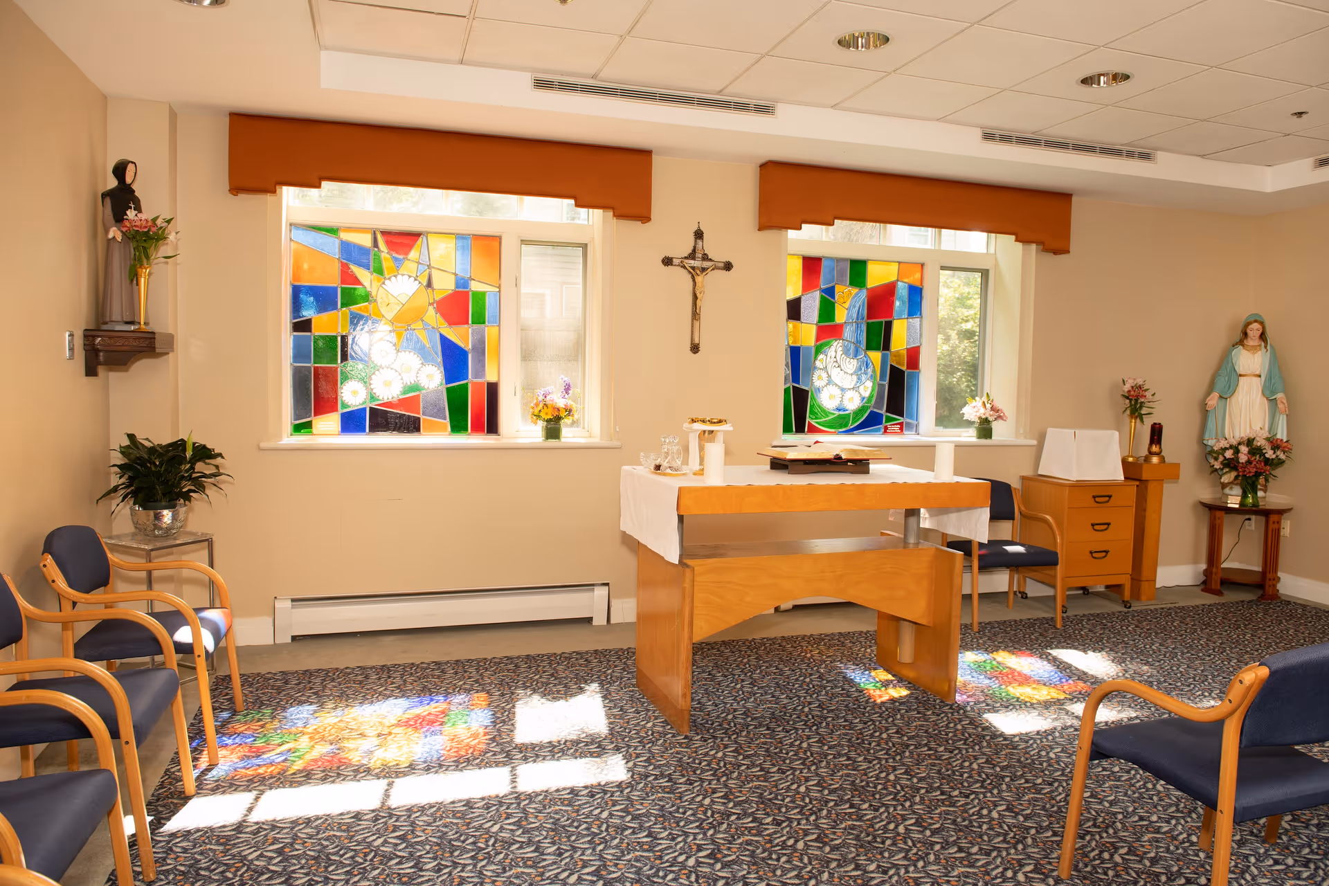 A small chapel room with stained glass windows casting colorful light patterns on the carpeted floor. The room features a wooden altar covered with a white cloth, a crucifix on the wall, religious statues, and several chairs arranged around the space.
