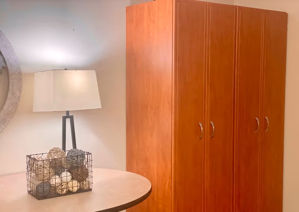 A corner of a room featuring a round table with a decorative wire basket filled with various textured balls, a modern table lamp with a rectangular white shade, and a large wooden wardrobe with four doors and silver handles against a light-colored wall.