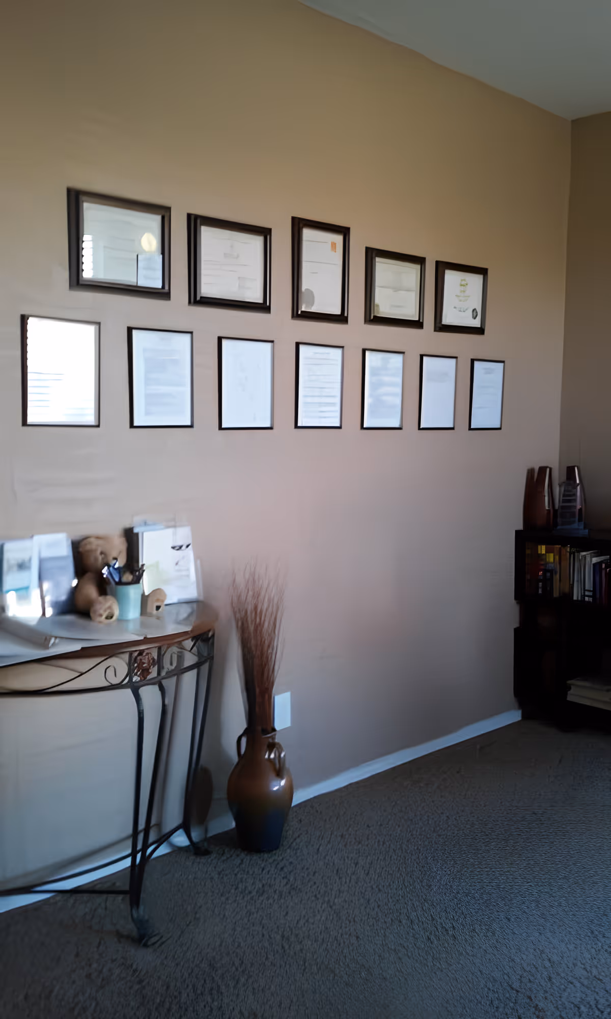 Interior corner of a room with a beige wall displaying two rows of framed certificates or documents. Below the frames is a decorative metal table with various items including a teddy bear and a cup with pens. Next to the table is a tall brown vase with dried branches. To the right, there is a dark wooden bookshelf with books and decorative items.