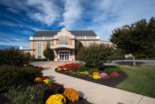 Front exterior view of a large brick building with a decorative entrance, surrounded by landscaped gardens with colorful flowers and trees under a partly cloudy blue sky.
