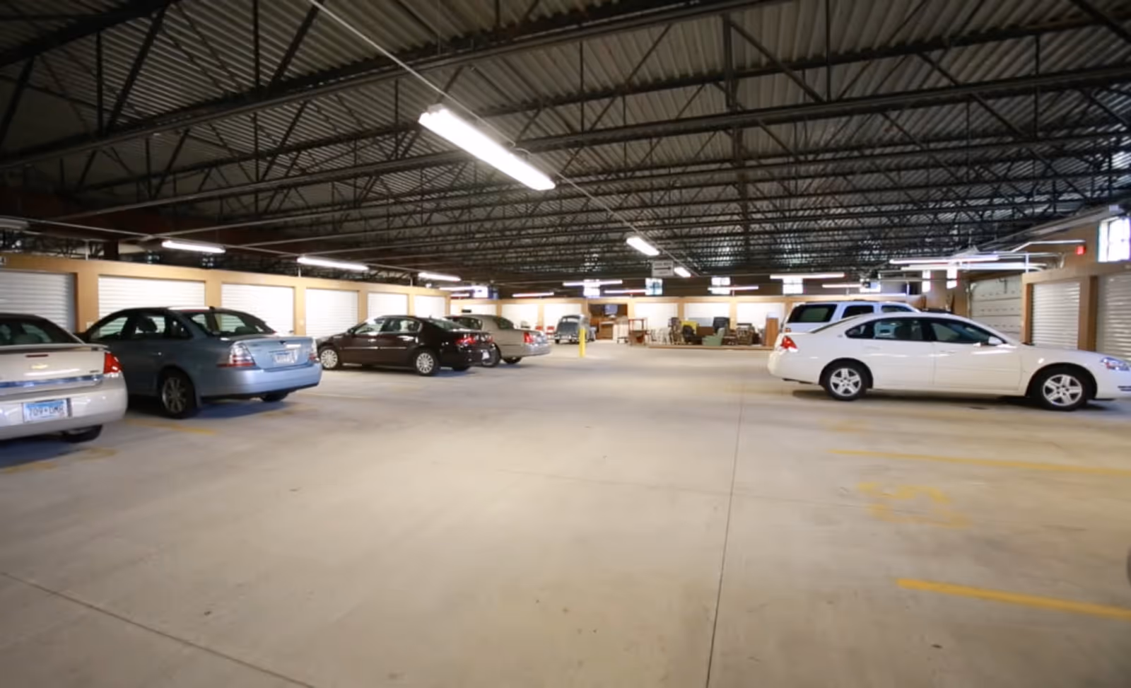 Indoor parking garage with several parked cars and storage units along the walls under a metal roof with fluorescent lighting.
