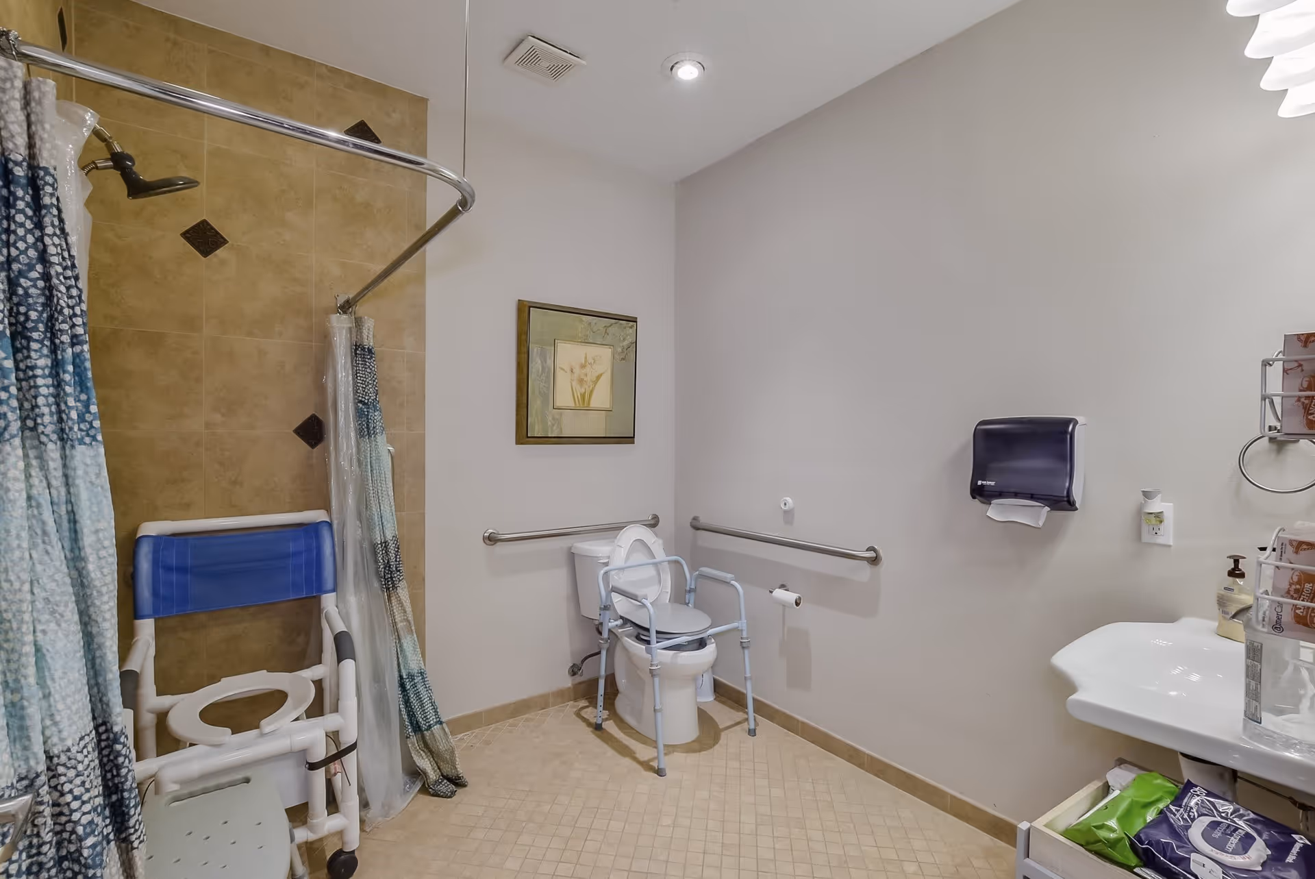 Accessible bathroom with a walk-in shower and shower chair, a toilet with grab bars and raised commode, sink, and paper towel dispenser.
