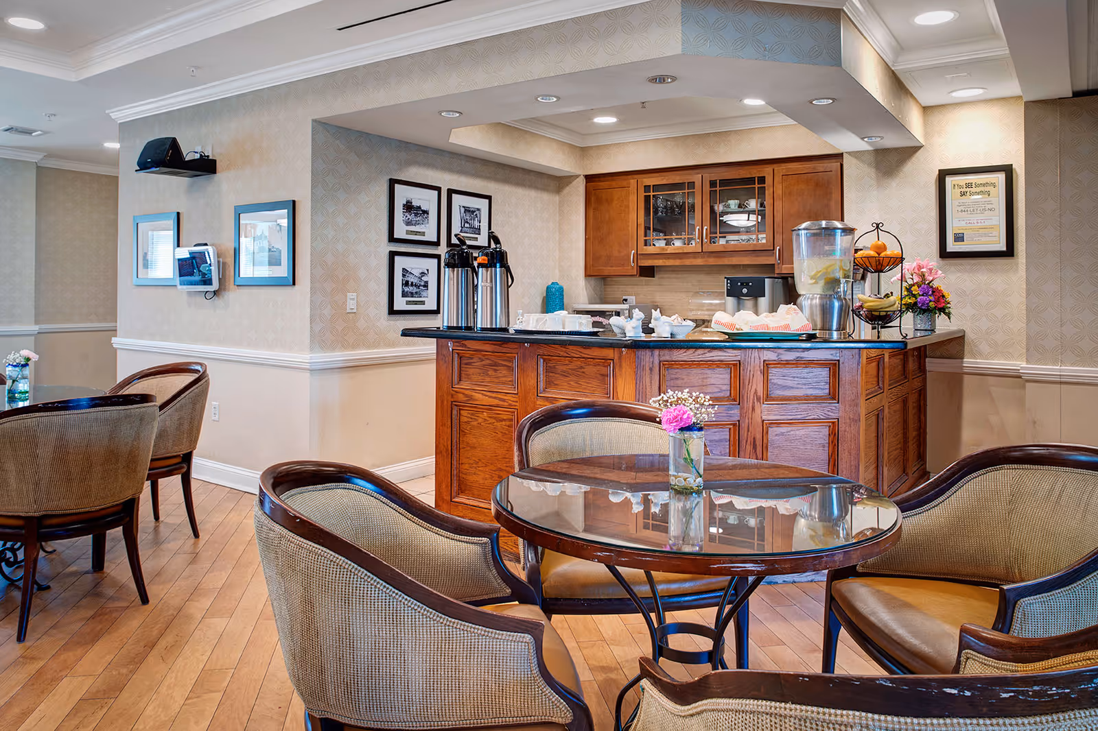 A cozy seating area with round glass-top tables and cushioned chairs in a senior living facility. In the background, there is a wooden counter with coffee dispensers, a water dispenser with lemon slices, a fruit basket, and a flower arrangement. The walls are decorated with framed pictures and patterned wallpaper.