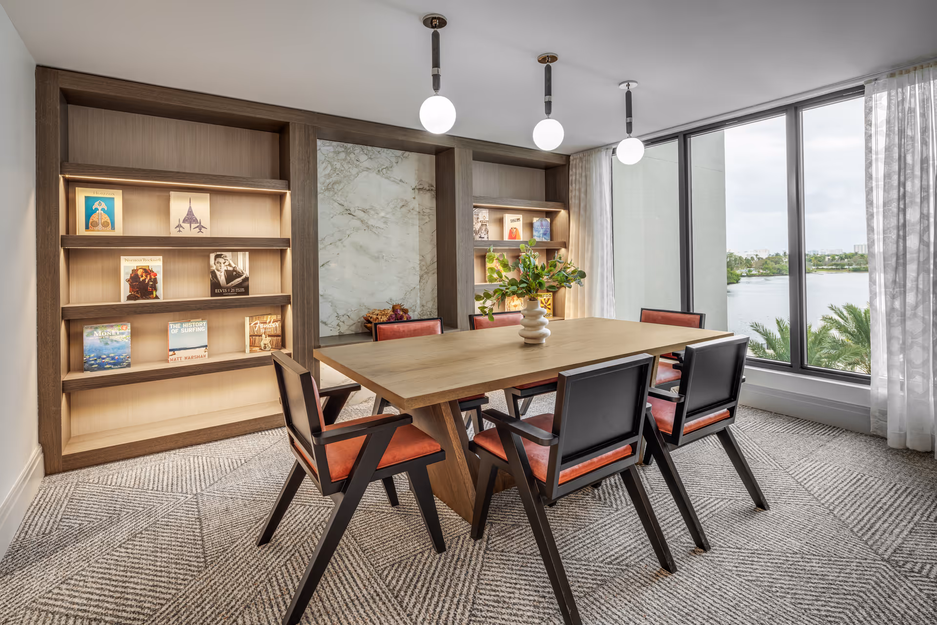 A modern dining room with a wooden table surrounded by six chairs with red cushions and black frames. Behind the table is a built-in wooden bookshelf with illuminated shelves displaying various books and decorative items. The room has large floor-to-ceiling windows with sheer white curtains, offering a view of a body of water and greenery outside. Three round pendant lights hang from the ceiling above the table.