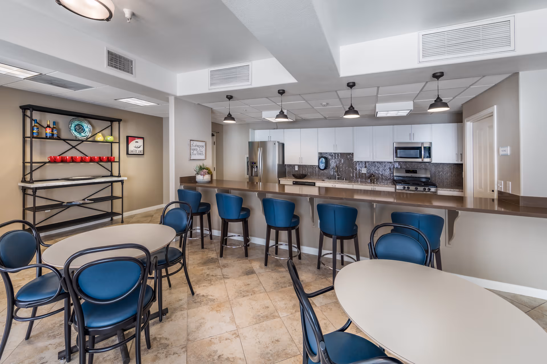 A modern communal kitchen and dining area with a long counter featuring six blue bar stools. There are two round tables with blue cushioned chairs. The kitchen has white cabinets, a stainless steel refrigerator, microwave, and stove. A black metal shelving unit with decorative items and red cups is against the wall. The floor is tiled, and the ceiling has recessed lighting and hanging pendant lights above the counter.