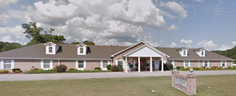Wide exterior view of Lynnfield Place, a single-story brick building with a pitched roof and dormer windows. The entrance features a covered porch with white columns, and there is a sign in front of the building on a brick base. The sky is partly cloudy.