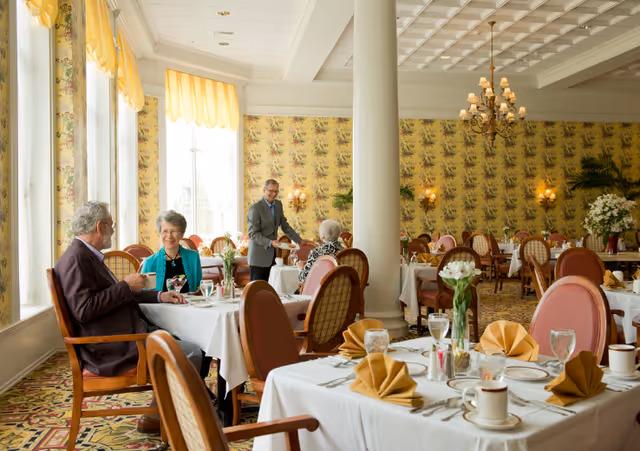Elegant dining room in a senior living community with tables set in white linens, folded napkins, chandeliers, and older adults being served.