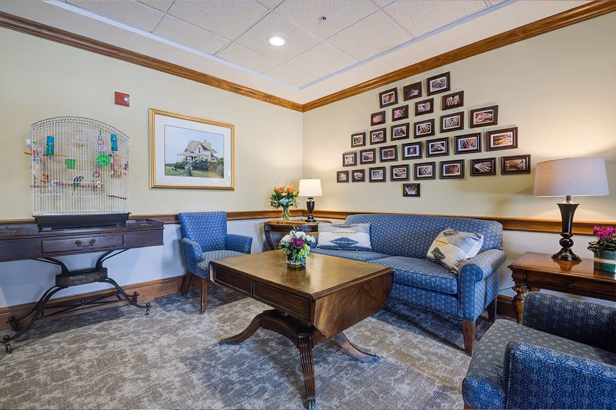 A cozy living room area with a blue patterned sofa, matching armchairs, a wooden coffee table, and side tables with lamps and flowers. On the left side, there is a birdcage on a wooden table. The walls are decorated with a framed picture of a house and a triangular arrangement of small framed photos.