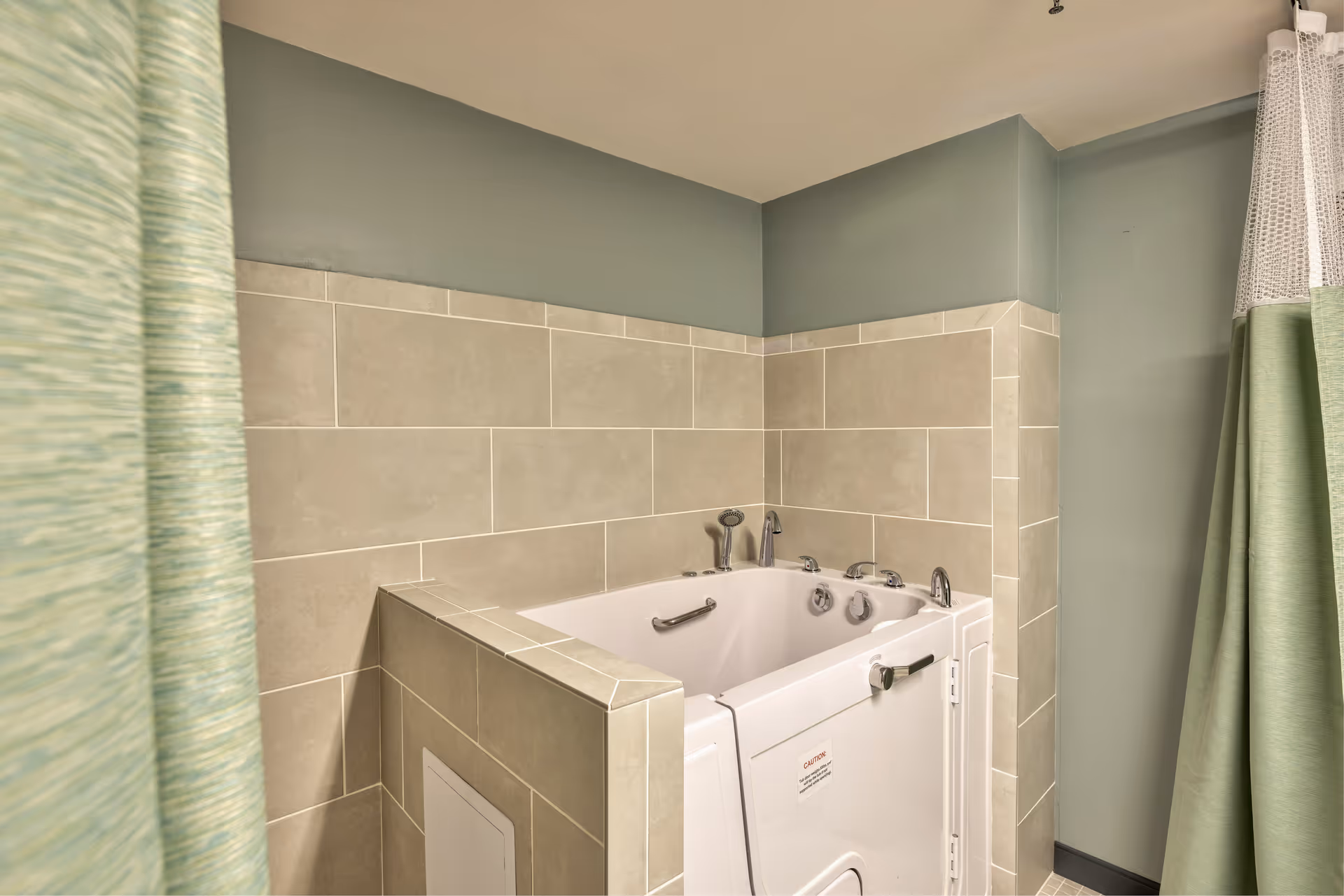 A walk-in bathtub with safety handles and multiple faucet controls, surrounded by beige tiled walls and green painted upper walls, partially enclosed by green curtains.