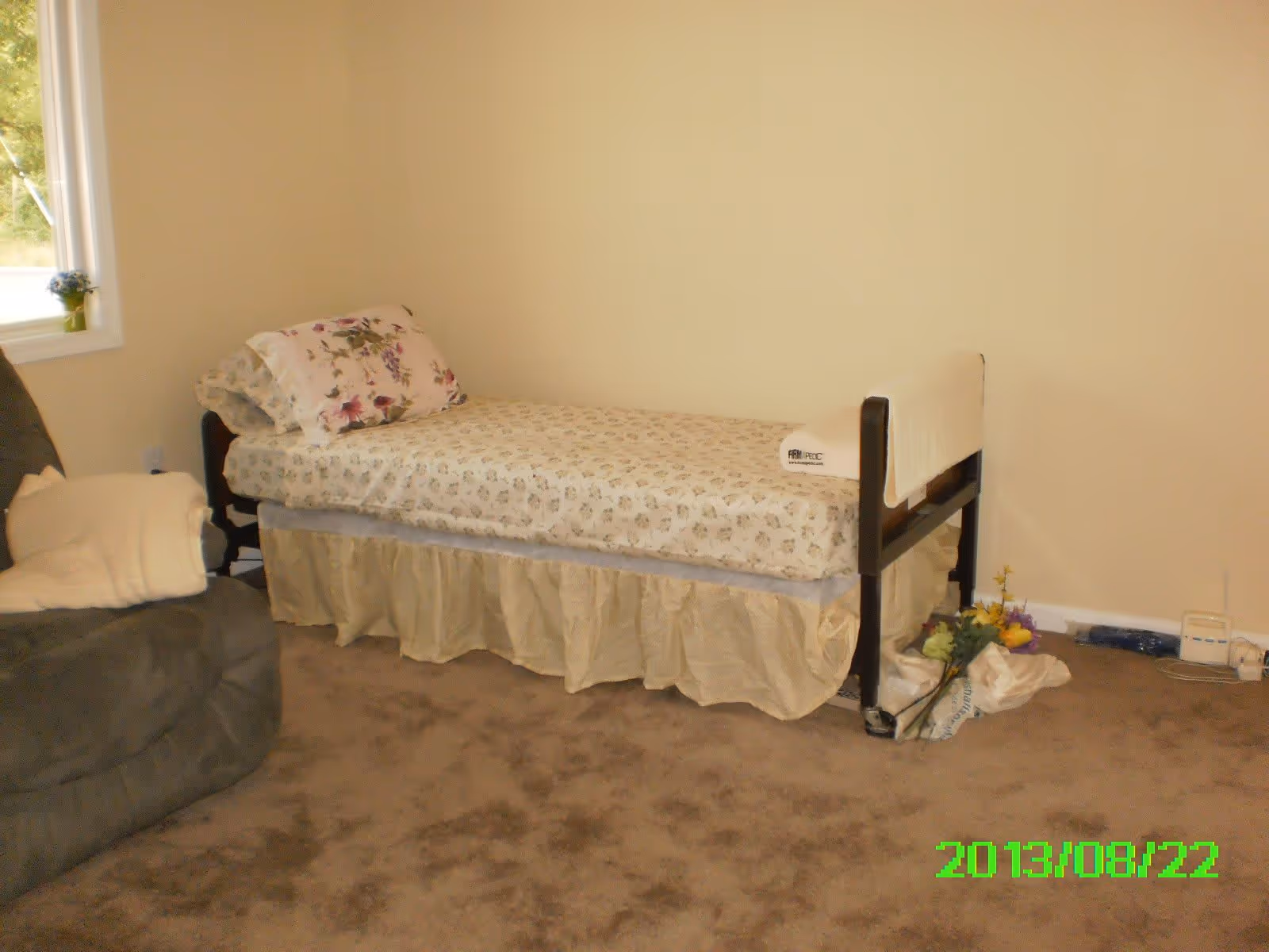 A simple bedroom with a single bed covered in floral bedding and a bed skirt. There is a floral pillow on the bed and a white towel draped over the bed's footboard. Next to the bed is a green armchair with a folded blanket on it. A small window with a potted plant on the sill is visible on the left wall. The floor is carpeted in brown, and there are some flowers and a plastic bag on the floor near the bed. The walls are painted a light beige color.