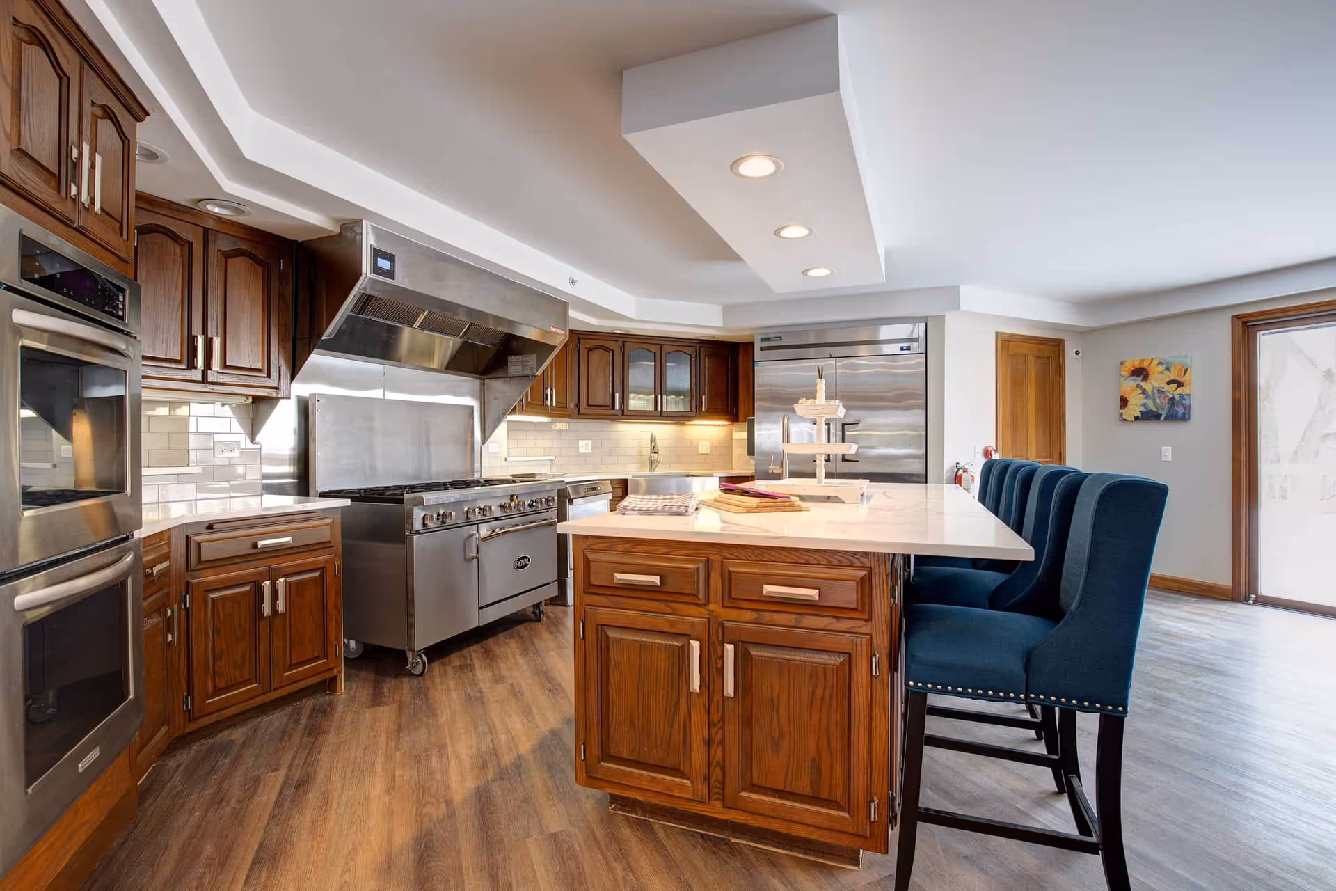 A spacious kitchen with wooden cabinets, stainless steel appliances including a large stove and double oven, a central island with a white countertop, and four blue upholstered chairs. The kitchen has wood flooring and a bright, clean atmosphere with a painting on the wall and a glass door leading outside.