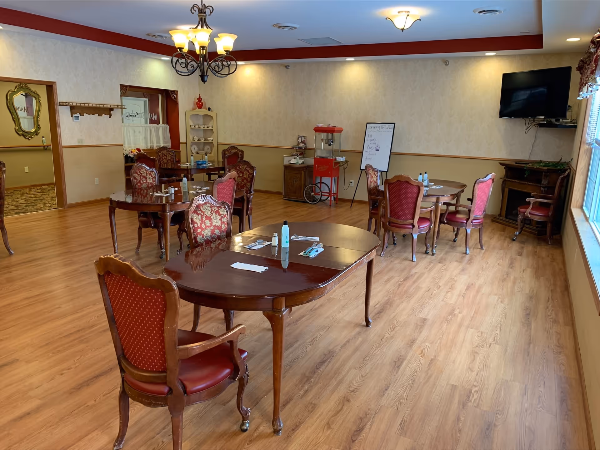 Spacious senior dining/activity room with wooden tables, upholstered chairs, a popcorn cart, whiteboard and wall-mounted TV.