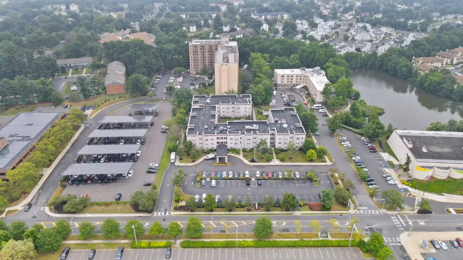 Aerial view of the Seaton Voorhees senior living facility showing multiple buildings, parking lots with covered parking, surrounding trees, and a nearby pond. Residential houses and roads are visible in the background.