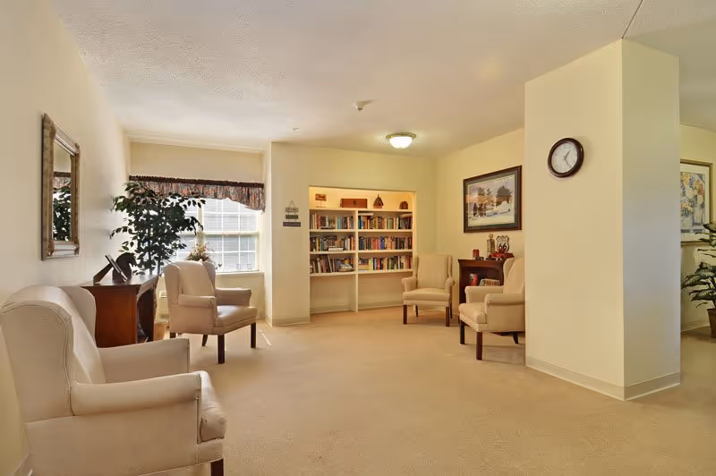 A cozy senior living common area with beige carpet and walls, featuring four beige armchairs arranged around a small wooden table. There is a built-in bookshelf filled with books, a framed picture on the wall, a clock, and a window with a valance letting in natural light. A potted plant and a mirror are also visible.