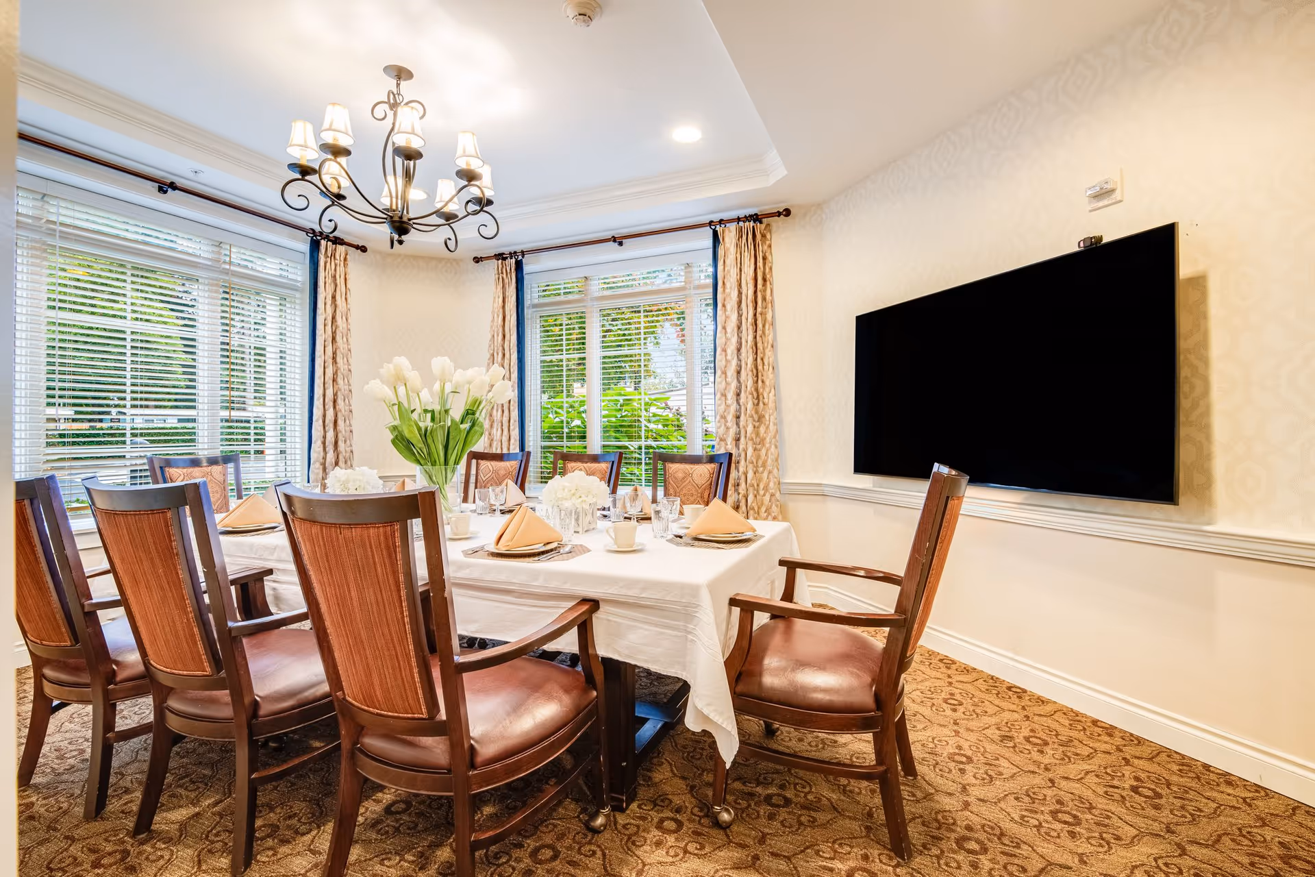 A well-lit dining room with a rectangular table covered with a white tablecloth, set with plates, glasses, napkins, and cups. The table is surrounded by eight wooden chairs with cushioned seats. A vase with white tulips and smaller floral arrangements decorate the table. Large windows with blinds and patterned curtains let in natural light. A chandelier hangs from the ceiling, and a large flat-screen TV is mounted on the wall.