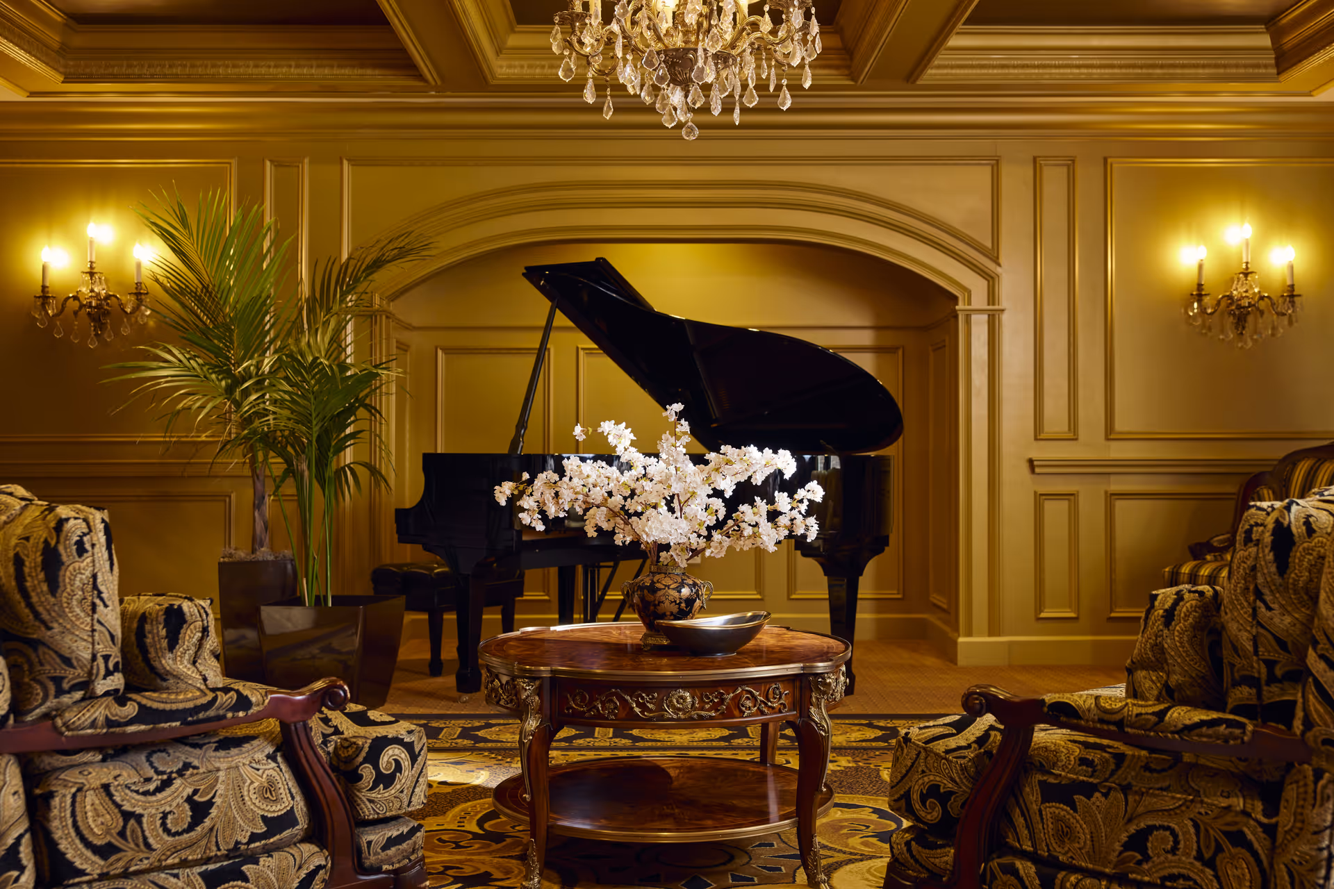 Elegant living room with ornate patterned armchairs, a round wooden coffee table with a floral arrangement and bowl, a grand piano in the background, a large potted plant, wall sconces with lit candles, and a crystal chandelier hanging from the ceiling.