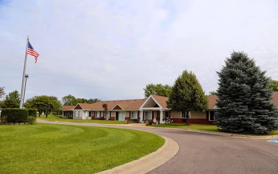 Exterior view of Regency Retirement Residence of Clinton showing a single-story building with a brown roof, surrounded by green grass, trees, and a curved driveway. An American flag is flying on a flagpole to the left.