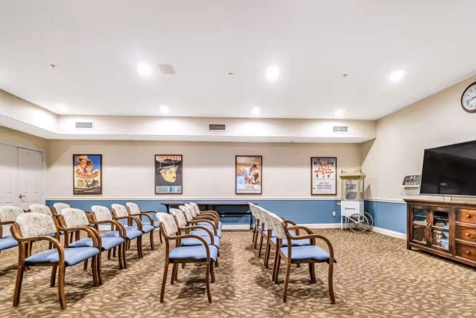 A room with two rows of wooden chairs with blue cushions facing a large flat-screen TV on a wooden cabinet. The walls are beige with a blue lower half and decorated with framed vintage movie posters. There is a popcorn machine in the corner and a clock on the wall above the TV.