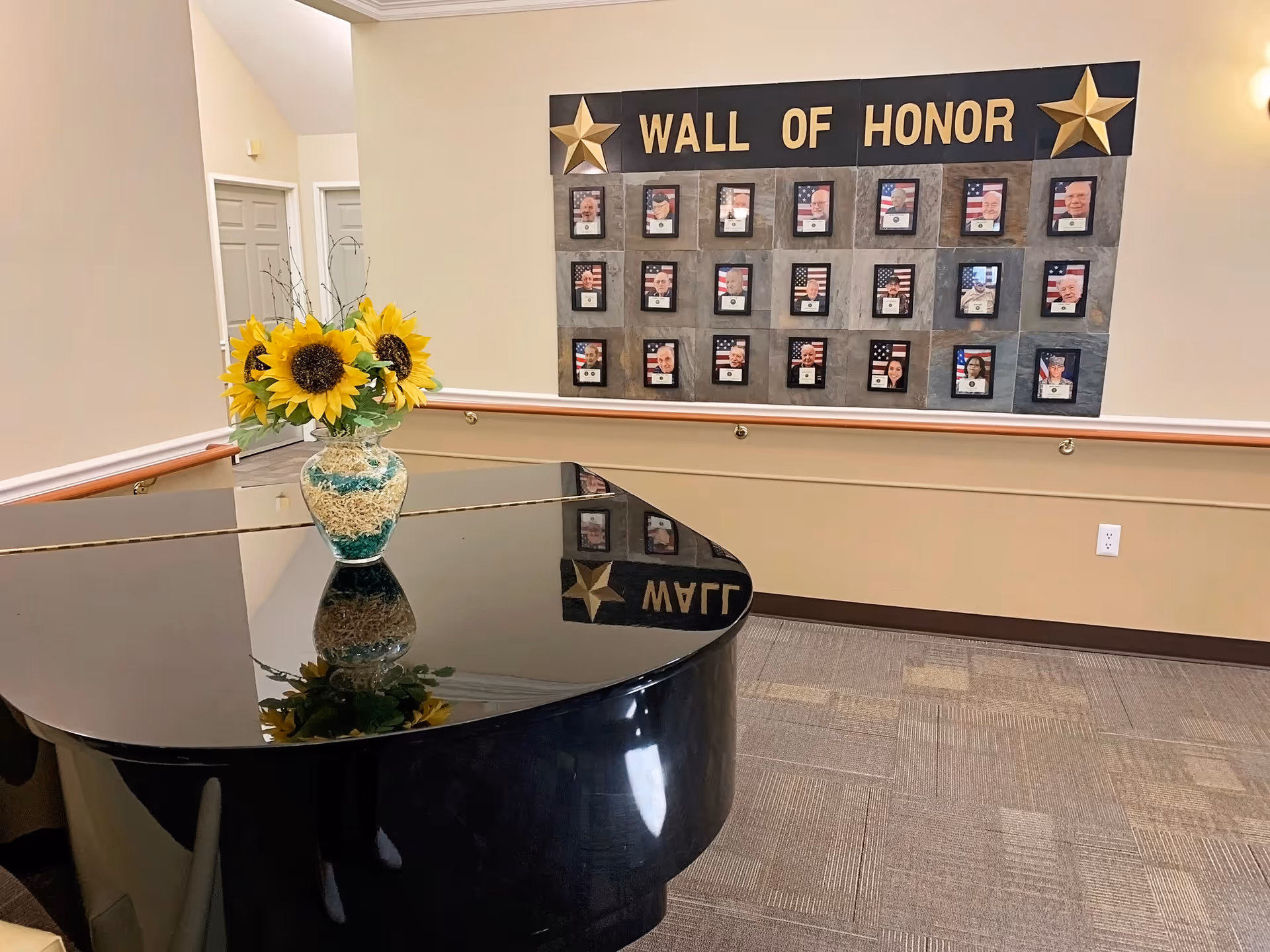 Interior view of a senior living facility hallway featuring a black grand piano with a vase of sunflowers on top. On the wall behind the piano is a 'Wall of Honor' display with framed photos and American flags, decorated with two gold stars.