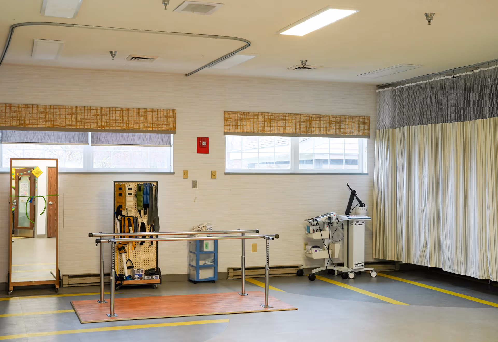 A physical therapy room with parallel bars on a wooden platform, a full-length mirror, a pegboard with therapy equipment, a small storage unit, and a medical device on a cart. The room has windows with woven blinds and a curtain divider on one side.