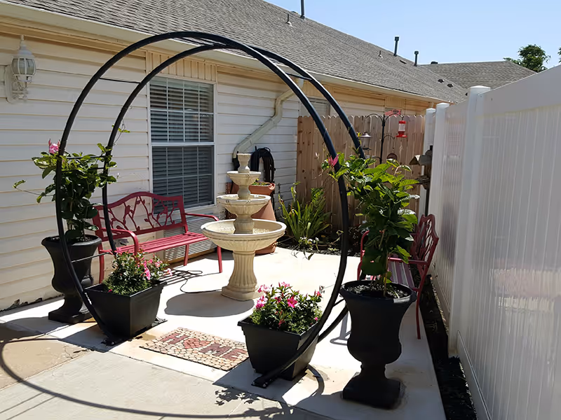 Outdoor patio area with a white three-tiered fountain in the center, surrounded by two black planters with green plants and pink flowers. There is a black metal arch over the fountain, two red benches against a beige building wall with a window, and a white fence on the right side. The ground is paved with concrete and there is a mosaic welcome mat in front of the fountain.