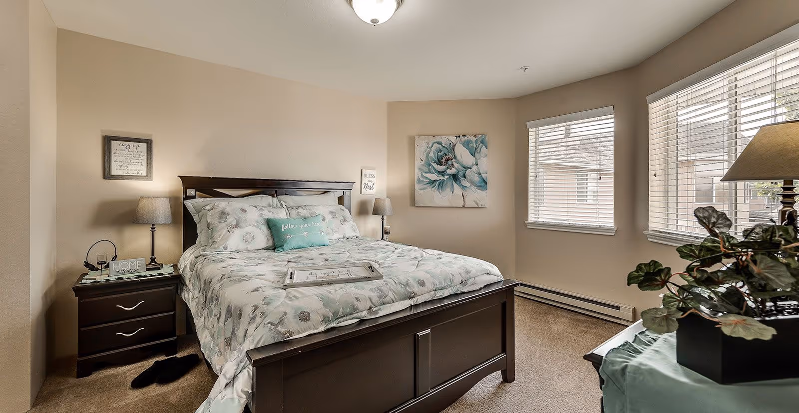 A cozy bedroom with a dark wooden bed frame and floral bedding. There are two bedside tables with lamps, a framed picture on the wall, and two windows with blinds letting in natural light. A potted plant is on a table near the windows.