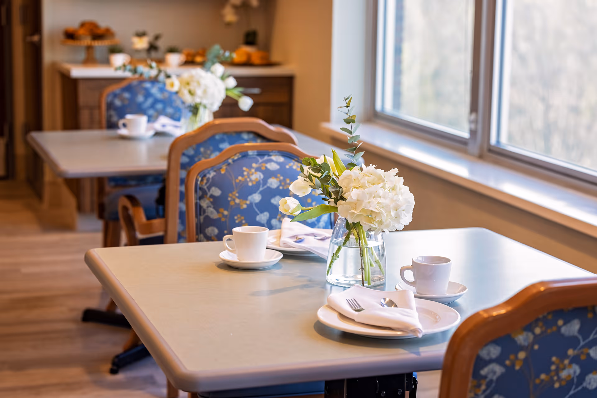 A dining area with tables set near large windows. Each table has a white floral centerpiece in a glass vase, white cups and saucers, plates with folded napkins, and silverware. The chairs have wooden frames and blue upholstery with a floral pattern.
