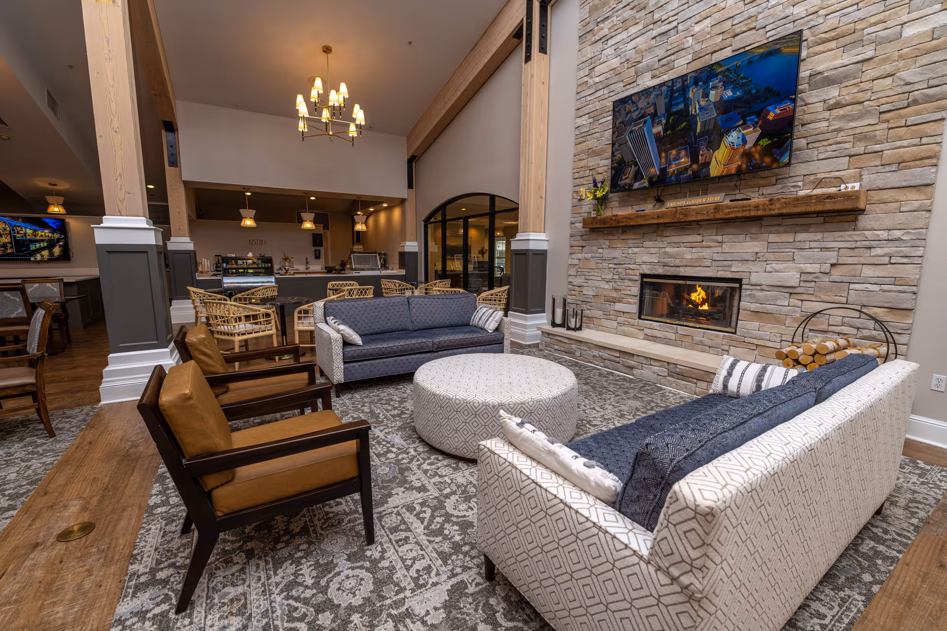 A cozy living room area with two patterned sofas, two brown armchairs, and a round ottoman arranged around a stone fireplace with a fire burning. Above the fireplace is a mounted flat-screen TV displaying a cityscape. The room features wooden beams, a patterned rug, and a view into a dining or kitchen area with wicker chairs and pendant lighting.