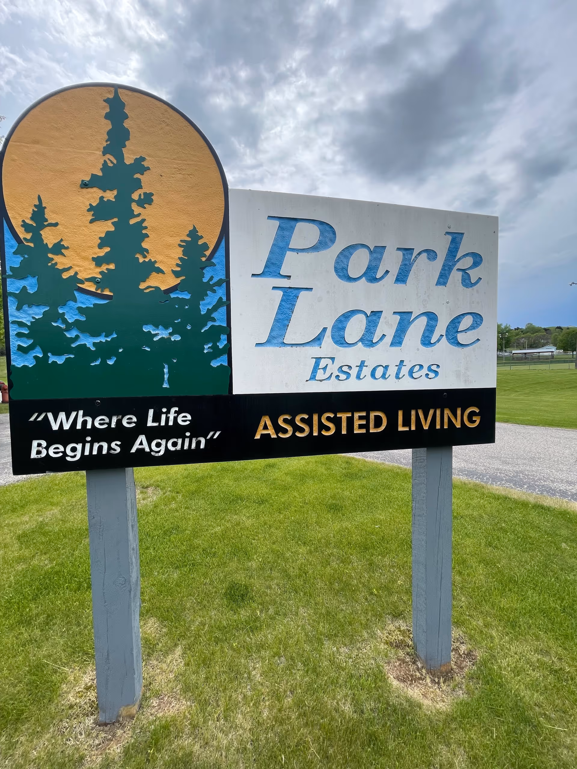 Outdoor sign for Park Lane Estates Assisted Living with a graphic of trees and a large orange sun or moon in the background, standing on a grassy area under a cloudy sky.