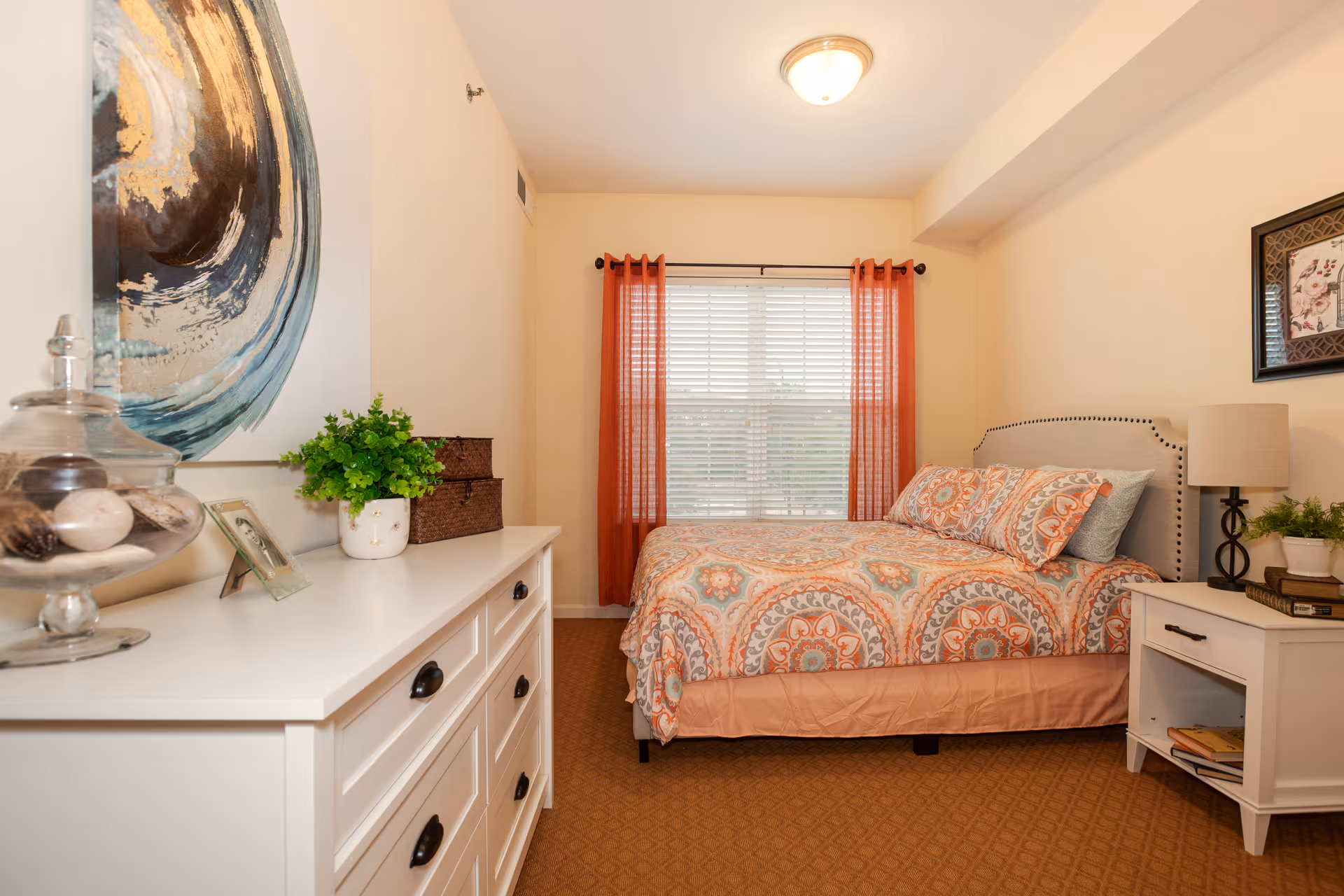 A cozy bedroom with a bed featuring a colorful patterned bedspread and matching pillows. The room has a white dresser with decorative items including a glass jar with seashells, a small plant, and a framed photo. There is a window with white blinds and orange curtains, a nightstand with a lamp, books, and a small plant. The walls are light-colored and decorated with framed artwork.