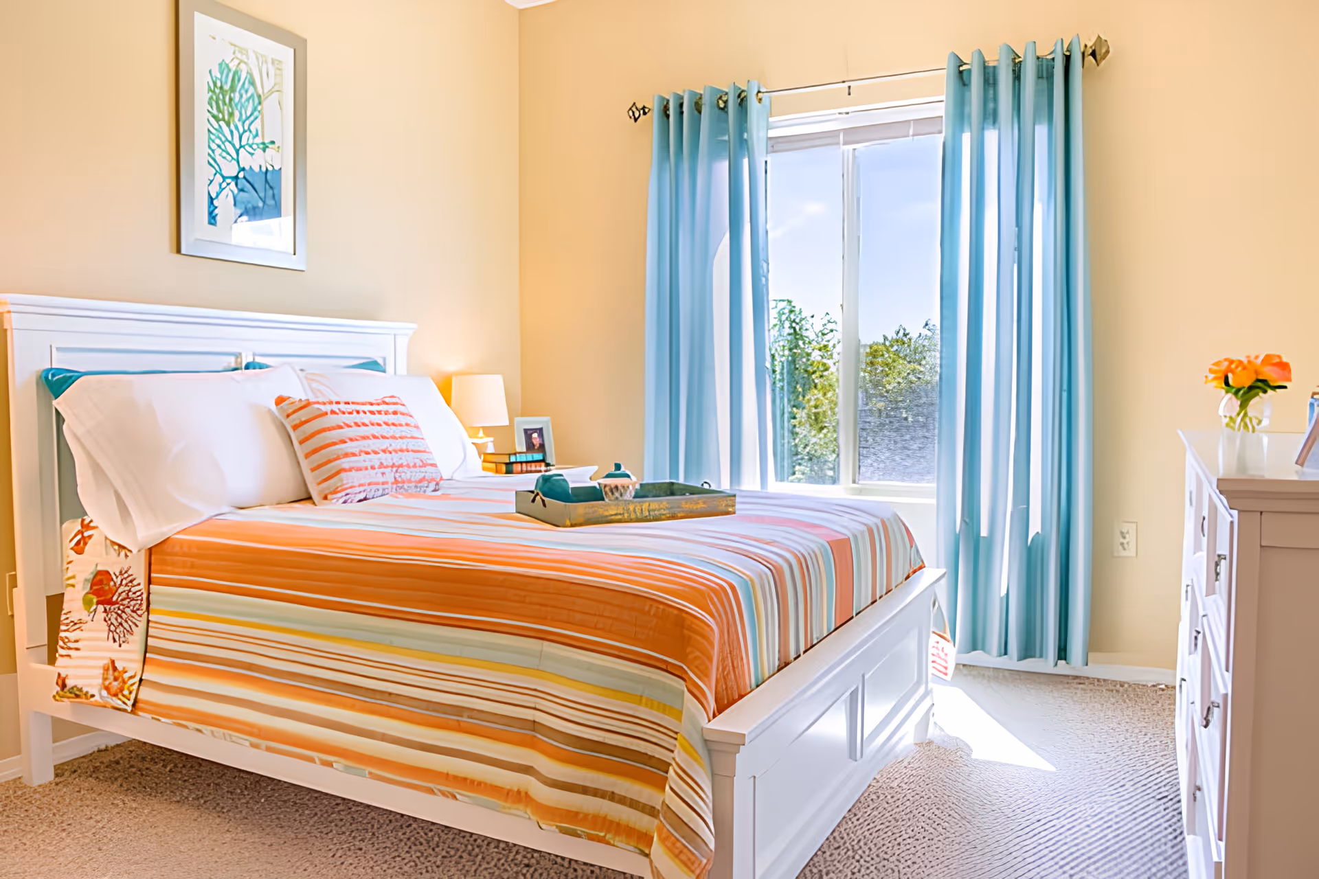Sunny bedroom with a white bed dressed in striped orange and teal bedding, blue curtains, and a window letting in natural light.