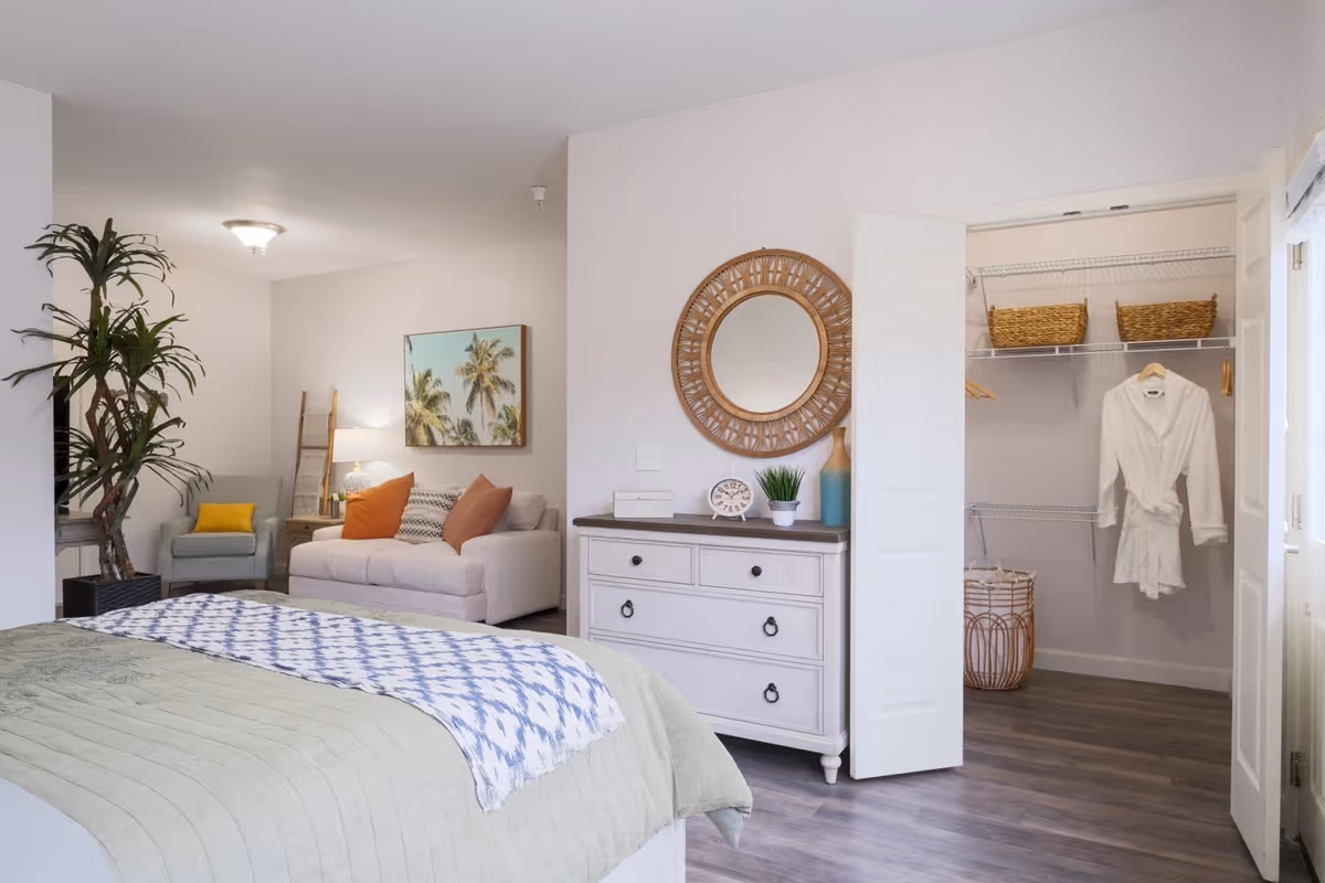 Bright furnished bedroom with a bed in the foreground, a dresser and open closet on the right, and a sitting area with a sofa and plant in the background.