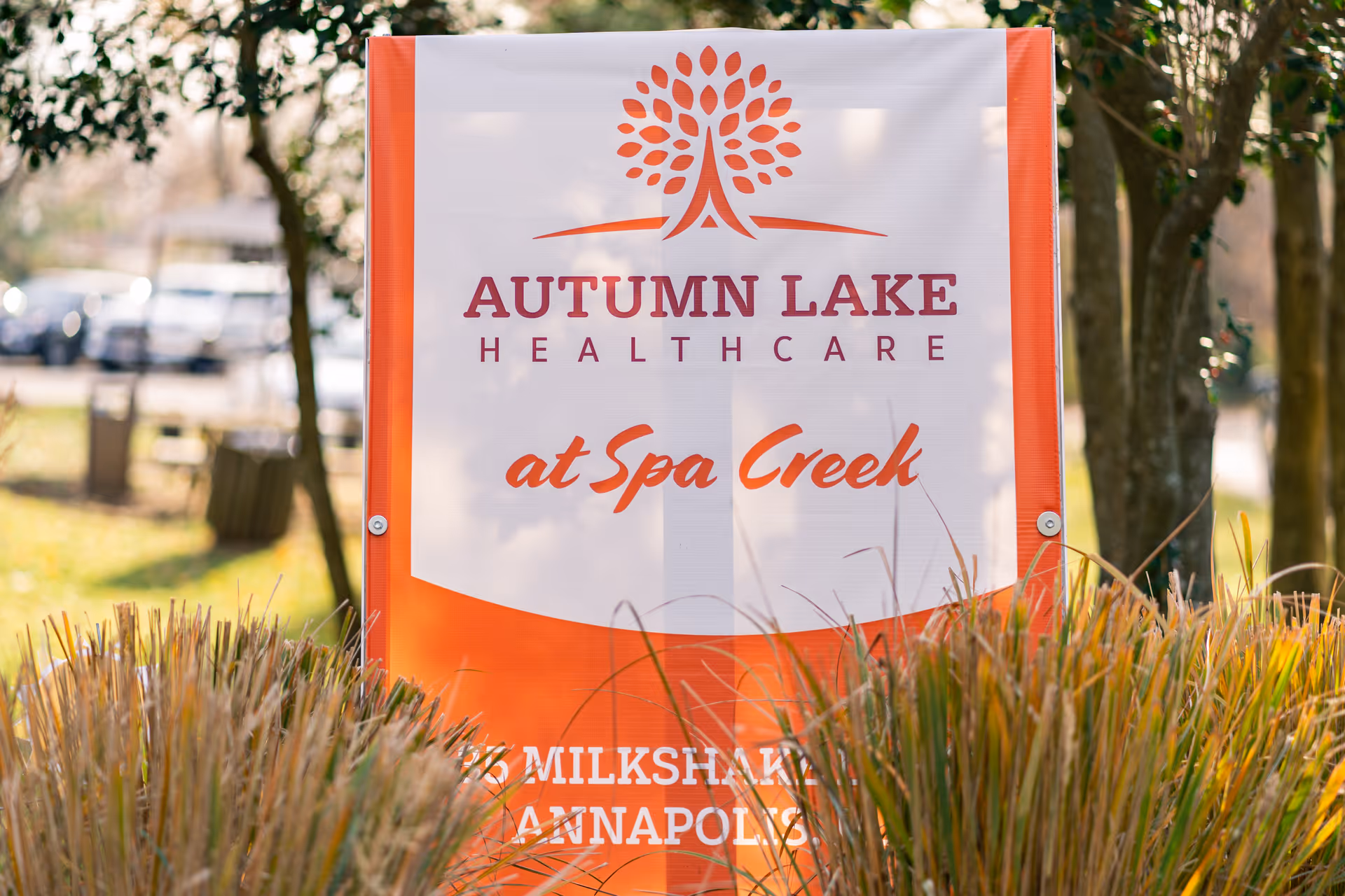 Outdoor sign for Autumn Lake Healthcare at Spa Creek surrounded by grass and trees, with a blurred background of parked cars and a bench.